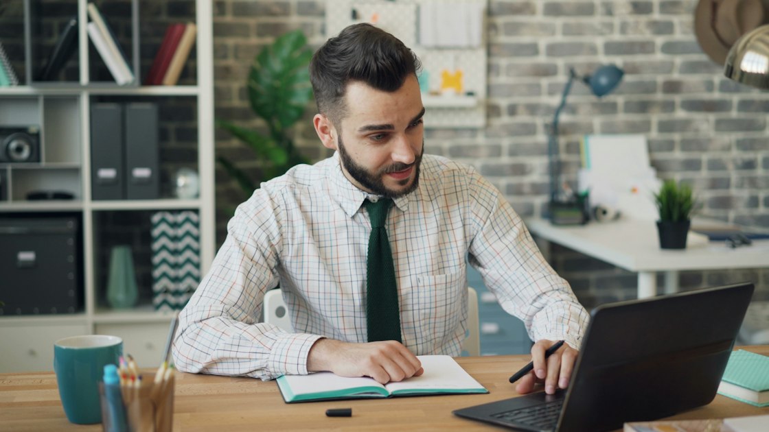 Bearded founder working on a laptop at a desk, taking notes in a notebook, illustrating the daily work of a solo founder building in public