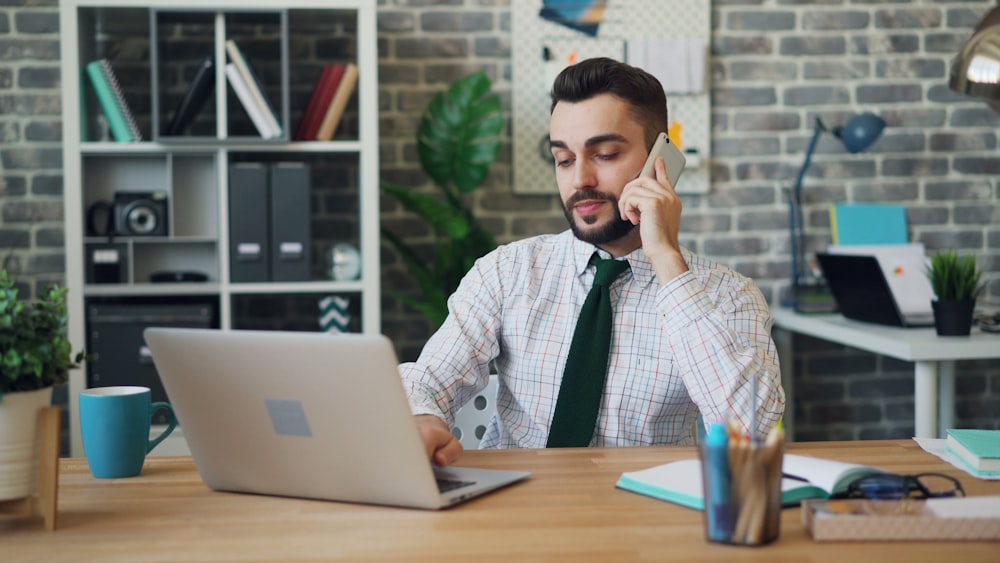 a man sitting in front of a laptop talking on a cell phone