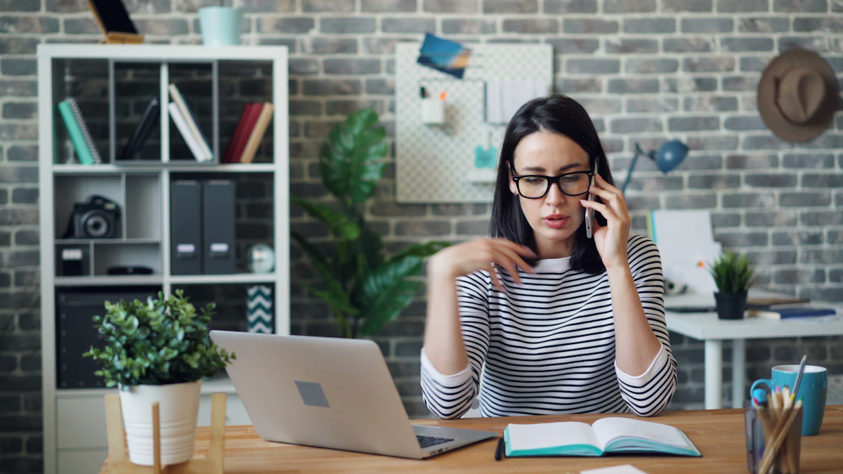 A woman in glasses takes a phone call at a wooden desk with an open laptop and notebook in a small office