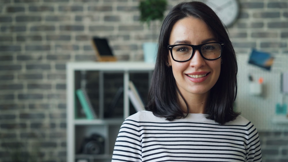 a woman wearing glasses standing in front of a brick wall