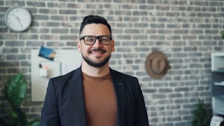a man wearing glasses standing in front of a brick wall