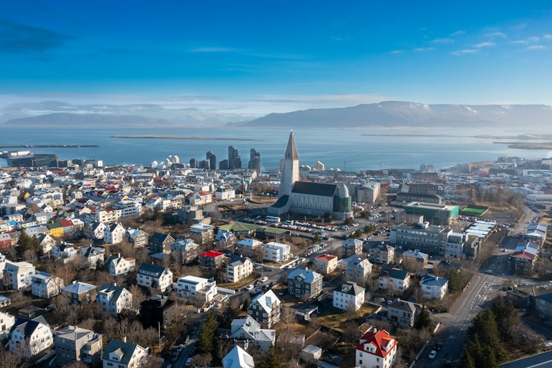 Aerial view of Reykjavík's colorful rooftops with Hallgrímskirkja's concrete spire rising above them and snow-streaked Mount Esja across the bay