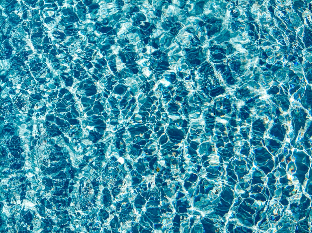 This image captures the sun-kissed surface of a clear, blue swimming pool. The water is shimmering with light, creating intricate patterns of ripples and reflections that give a sense of movement and tranquility.