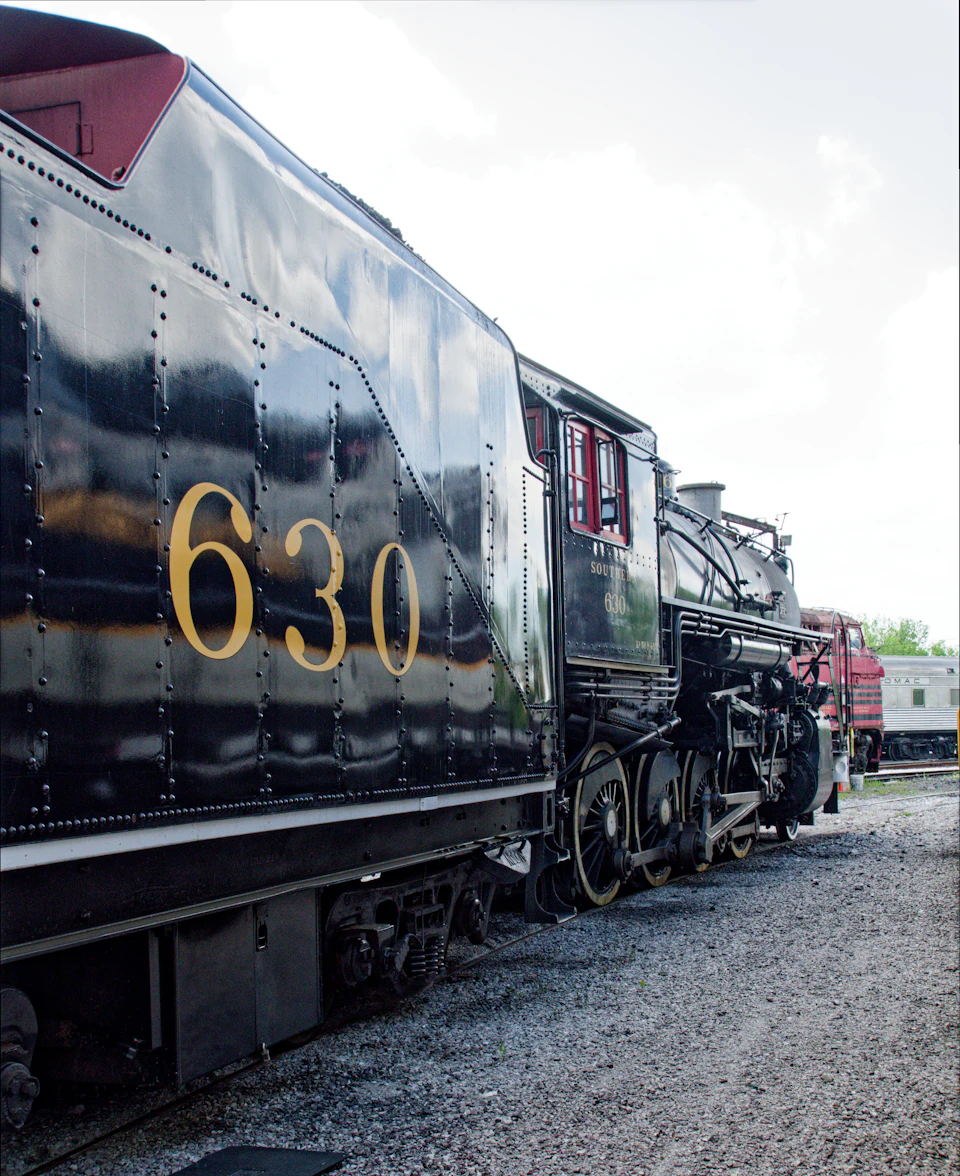 The image shows a black and yellow steam locomotive numbered 630, parked on a gravel lot with other trains visible in the background. The locomotive is facing towards the right side of the frame, giving a clear view of its design and features.
