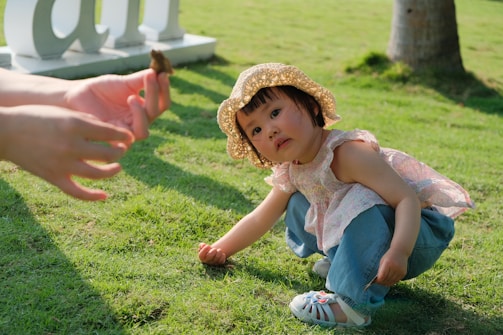 a little girl sitting on the grass with a hand reaching for a piece of food