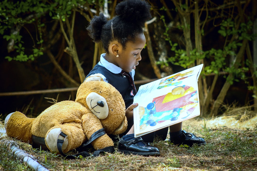 a young girl sitting on the ground reading a book to a teddy bear