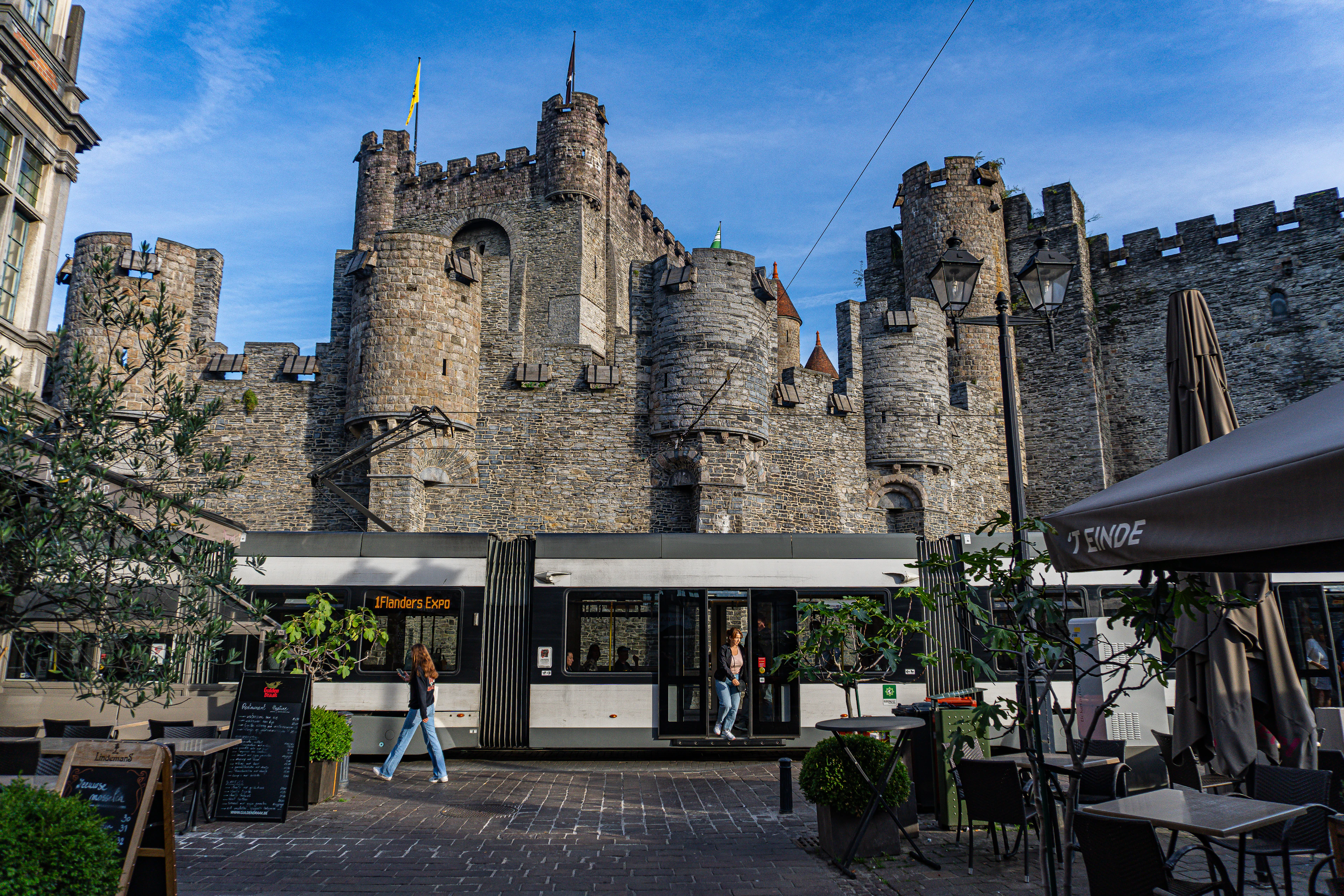 Gravensteen's grey stone towers rise over an outdoor cafe terrace under a blue sky.