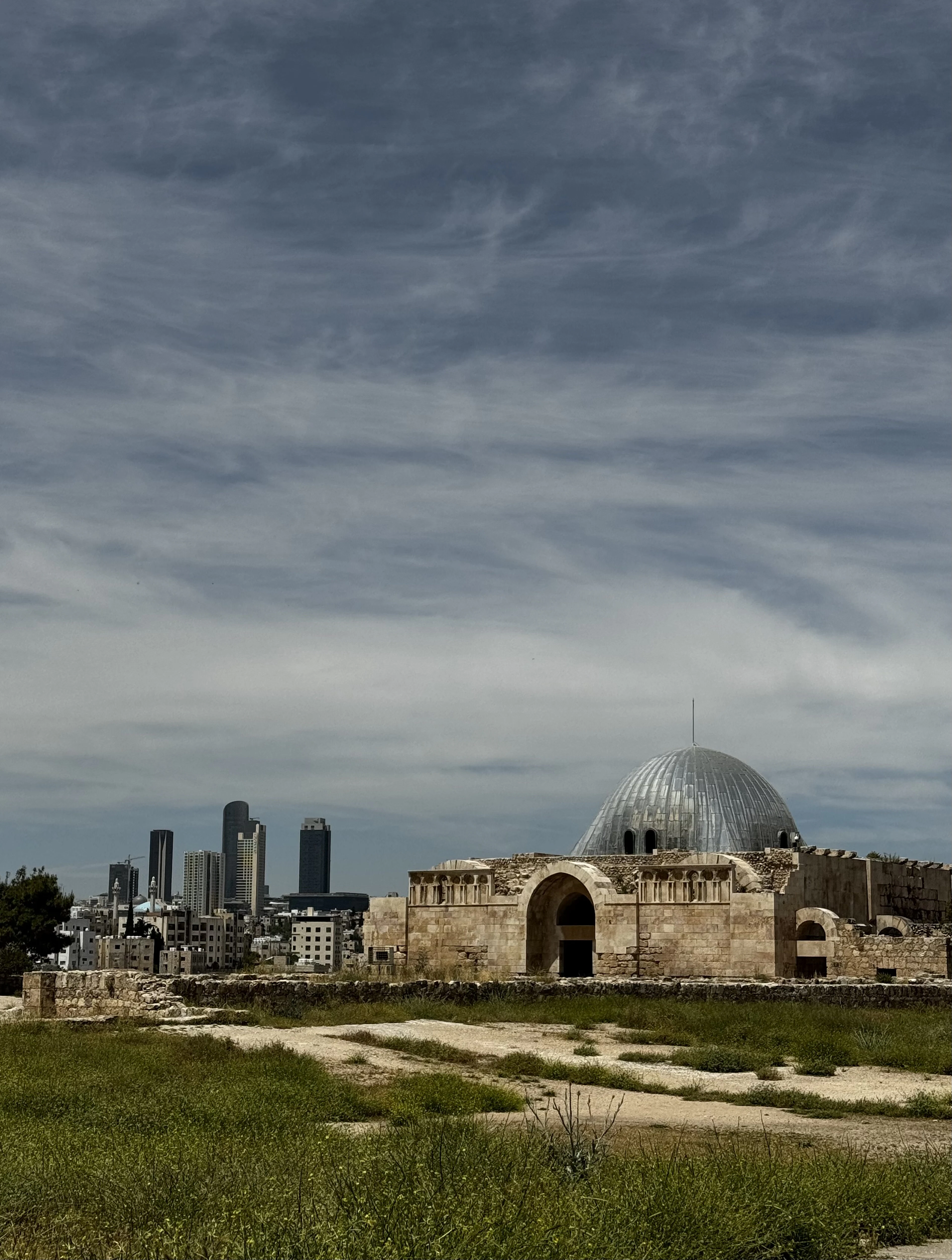 The Citadel's ancient dome sits before Amman's modern high-rise skyline under a moody sky.