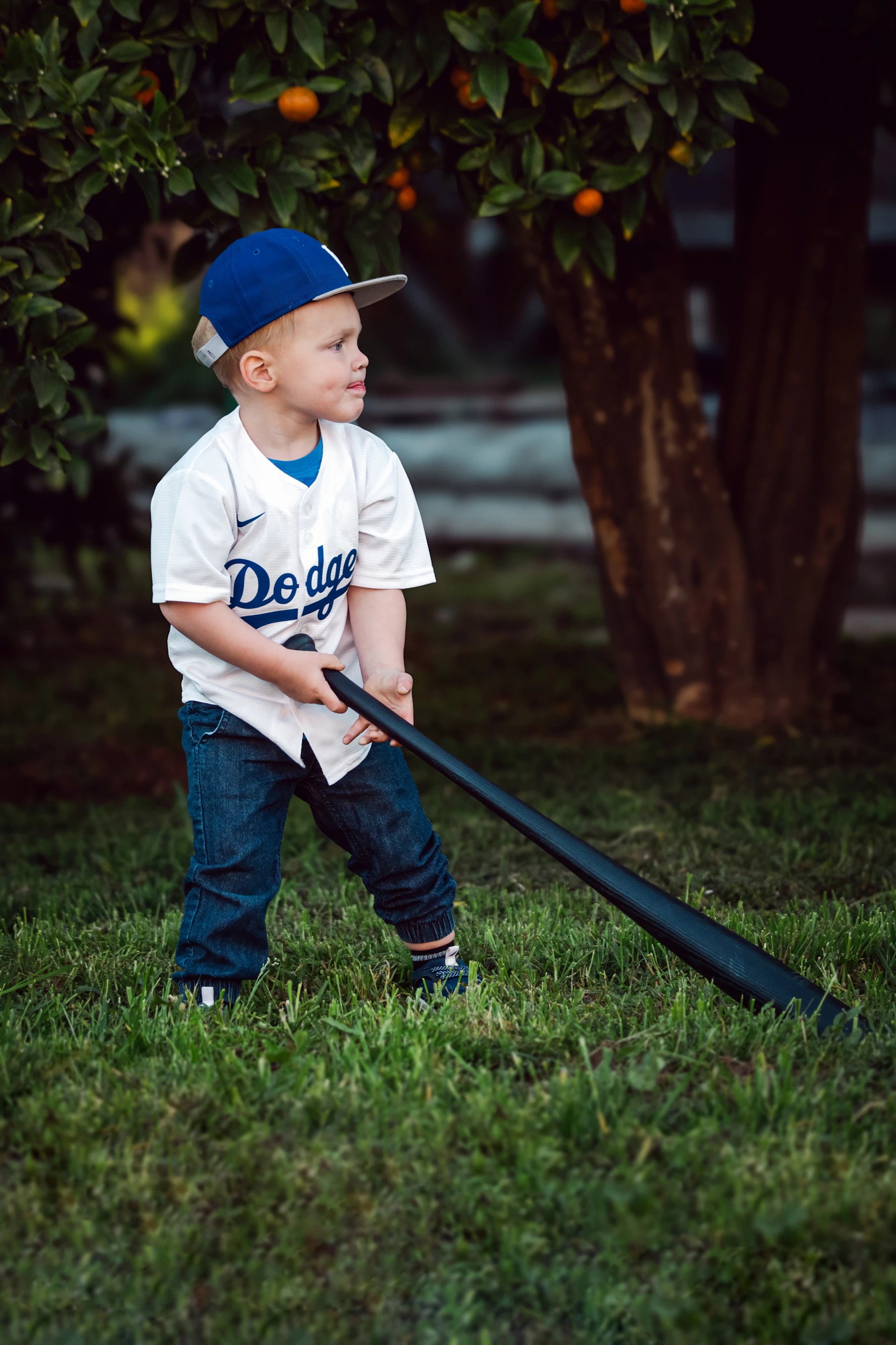 A small boy in a ball cap and white jersey holding a thin plastic bat under a backyard tree