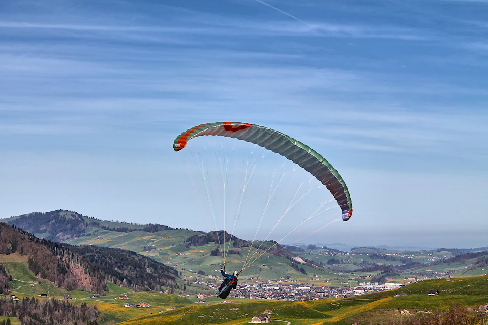The image depicts a person paragliding over a verdant valley, with their body suspended in mid-air and attached to a parachute that is spread out behind them. The parachute has a striking combination of red and green colors against the clear blue sky. Below the parachute flyer, there are several buildings nestled among the hills, suggesting a populated area at a lower altitude.