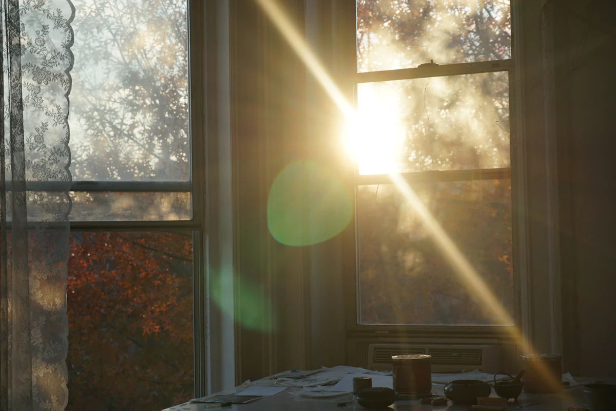 Lumière du matin traversant une fenêtre à rideaux, feuillage d'automne visible dehors, table dressée en contre-jour