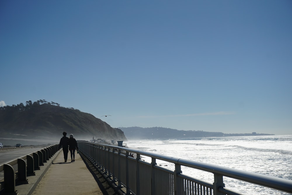 a couple of people walking down a sidewalk next to the ocean