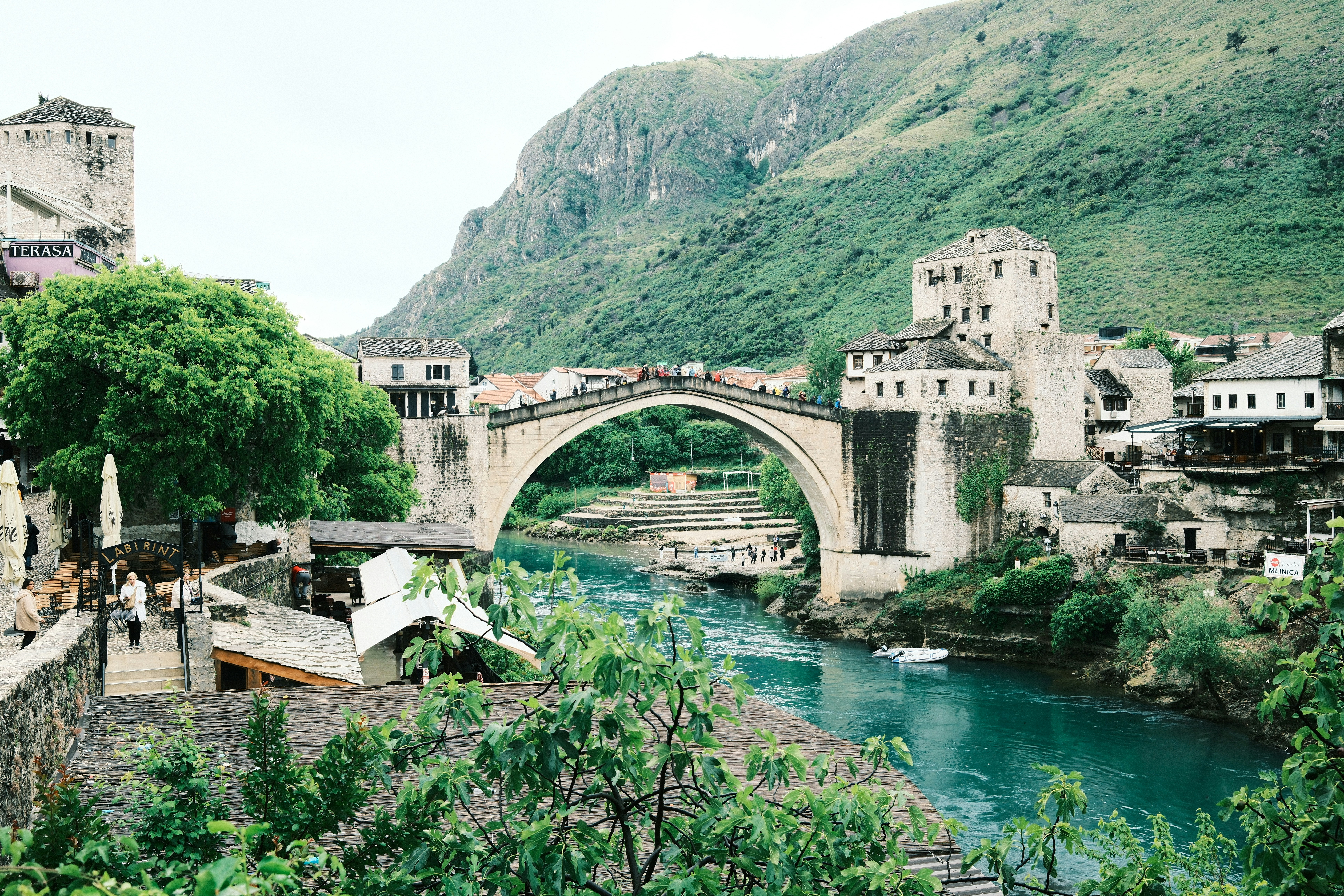 a river flowing under a bridge next to a lush green hillside, Mostar