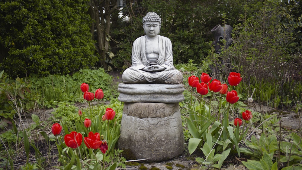 a buddha statue sitting on top of a stone statue surrounded by red flowers