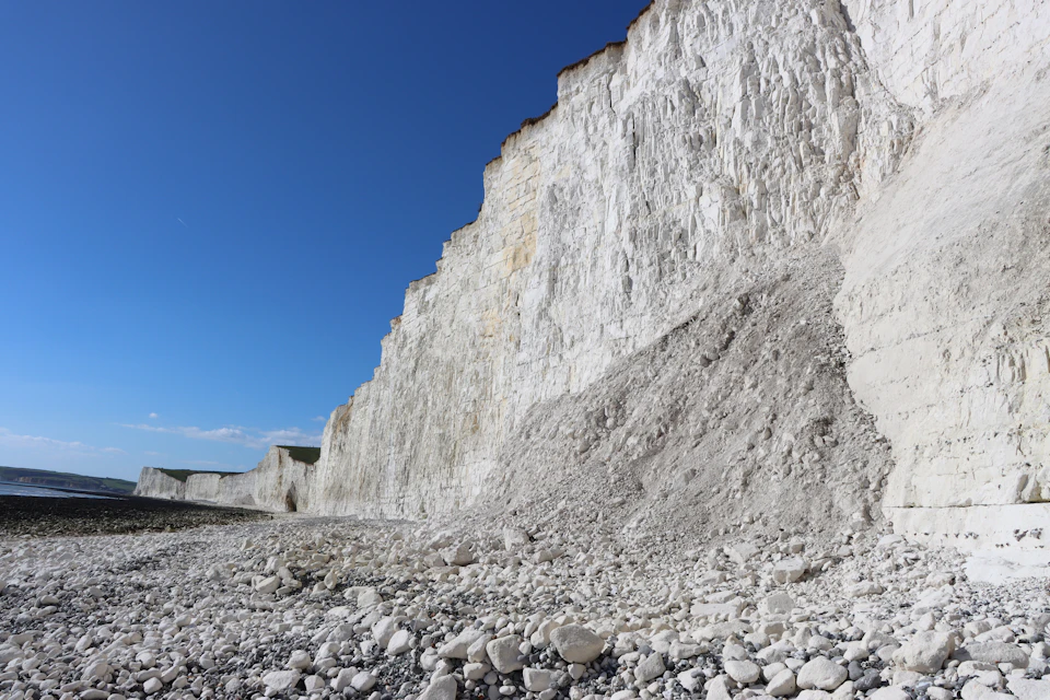 The image depicts a dramatic cliff face with white rock formations, some of which have been eroded and are now covered in grayish-white sediment. The cliff is situated on a beach, where small rocks can be seen scattered around it. The sky above the scene is clear blue, providing a striking contrast to the ruggedness of the cliff face.