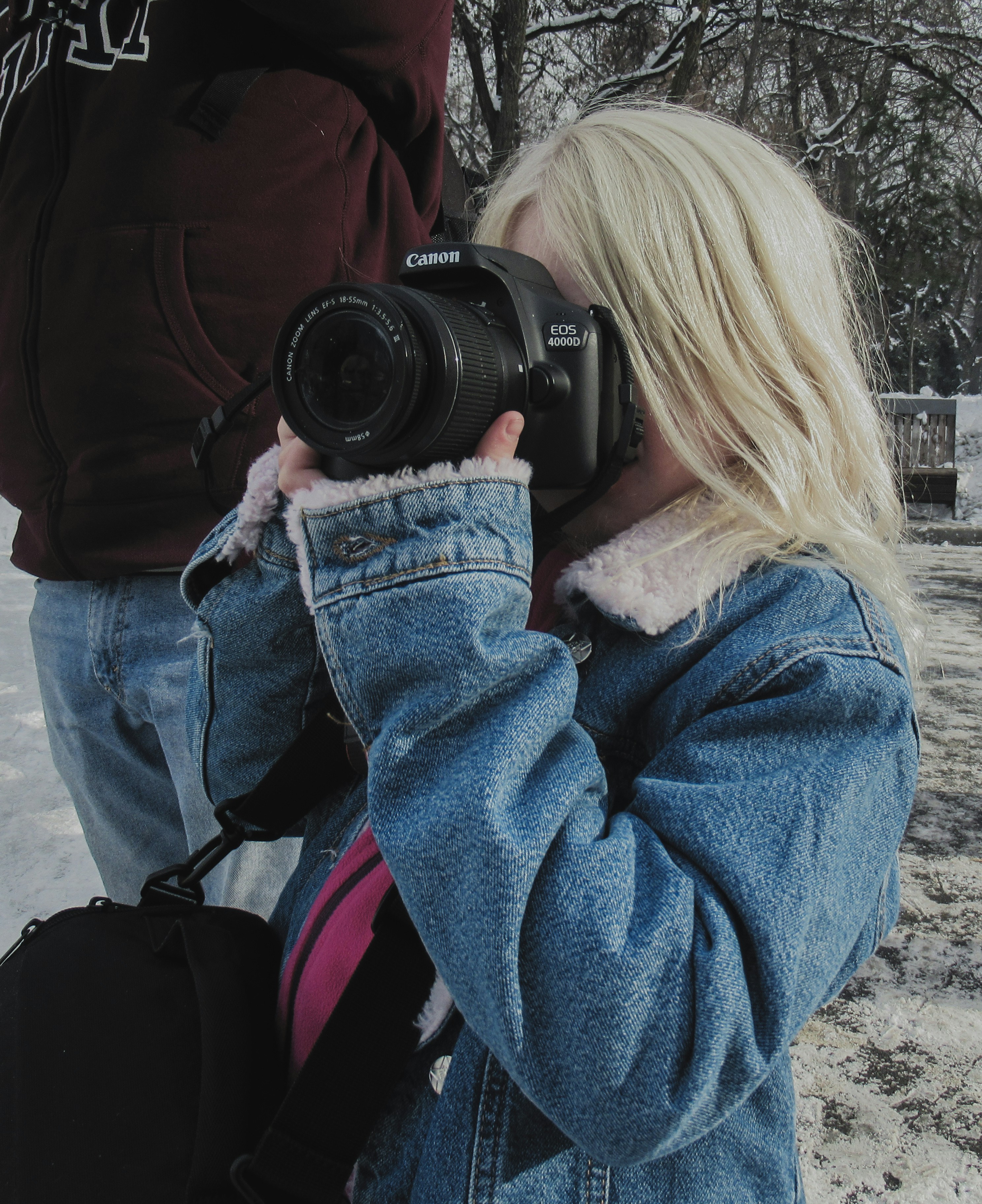 Photograph shows a young photographer in a denim jacket using a Canon camera outdoors in a snowy setting.