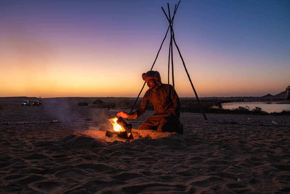 a man sitting next to a fire on top of a sandy beach
