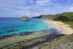 a sandy beach with clear blue water and green hills in the background