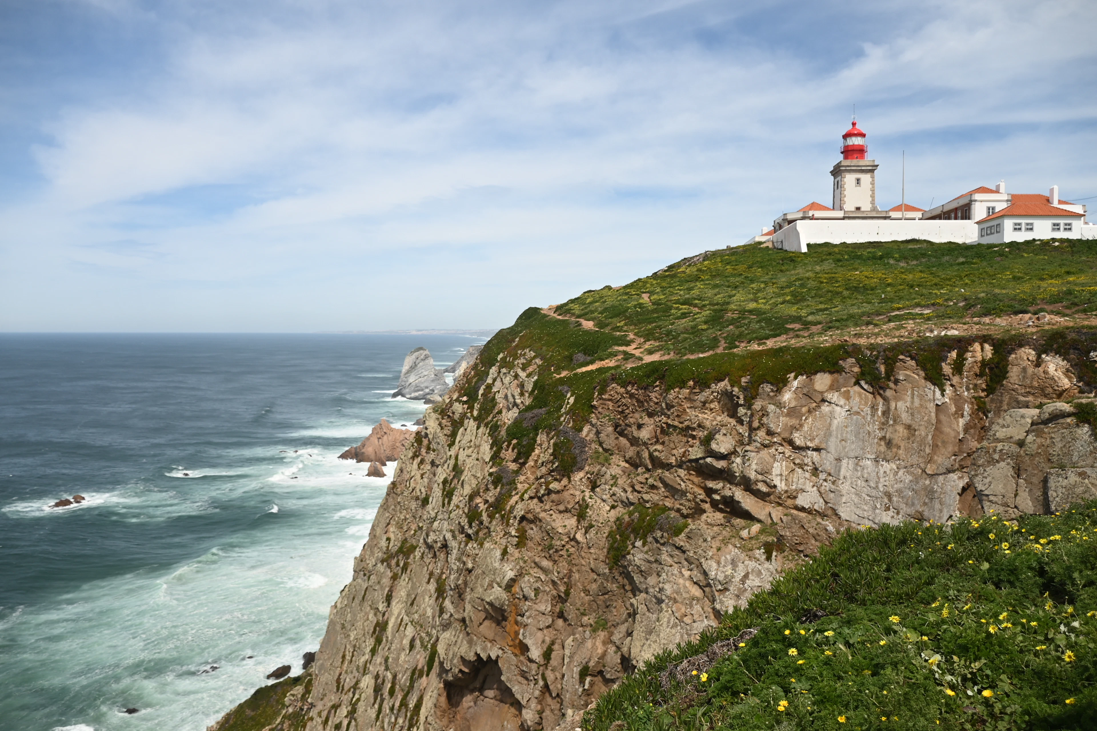 The red-capped lighthouse of Cabo da Roca standing on the grassy clifftop at the westernmost point of mainland Europe, the Atlantic breaking white on the rocks far below and yellow wildflowers in the foreground.