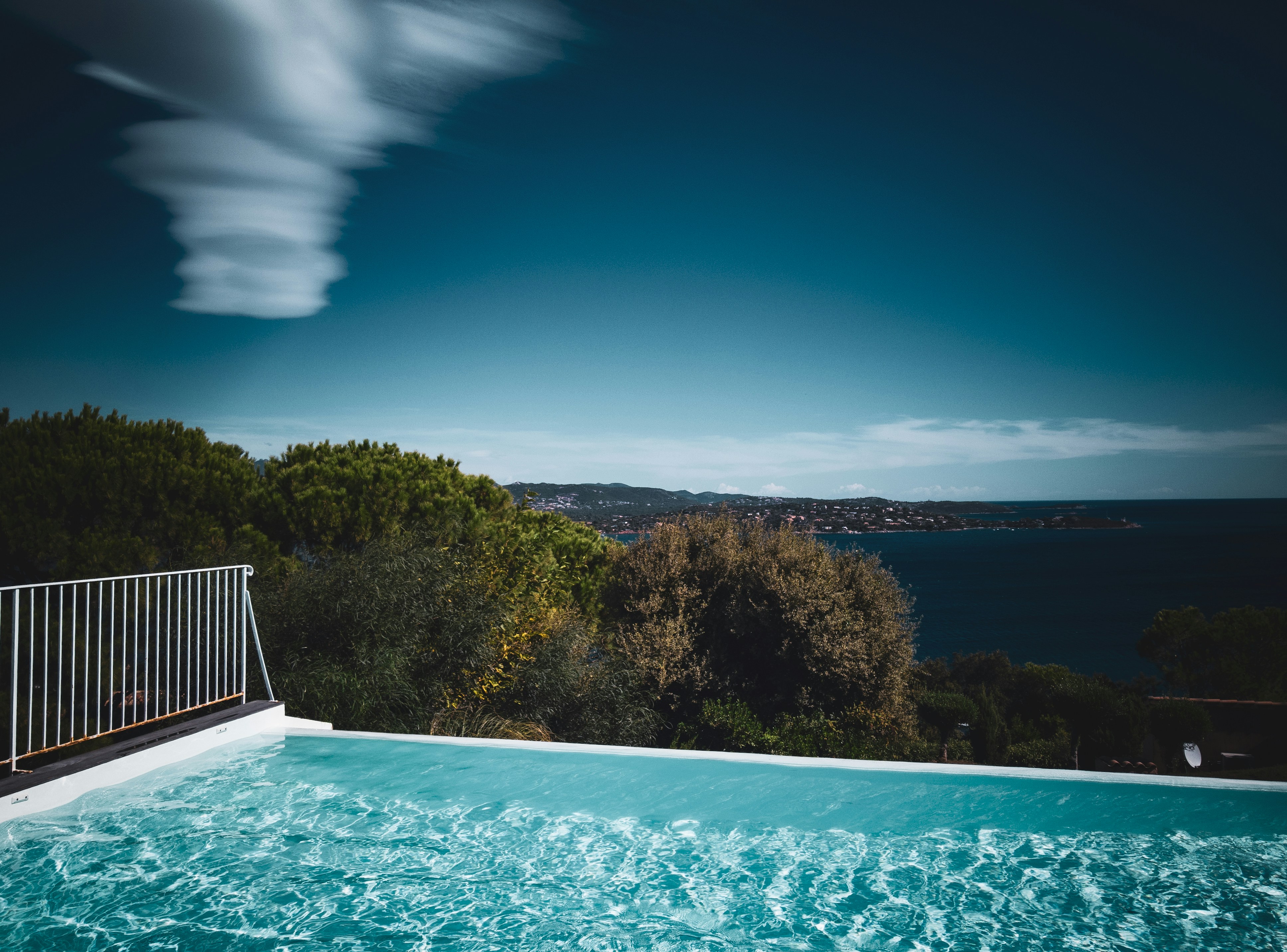 a swimming pool with a view of the ocean, An infinity swimming pool with a view of the sea, featuring a clear blue sky with streaky clouds above and dense greenery surrounding the area. The pool