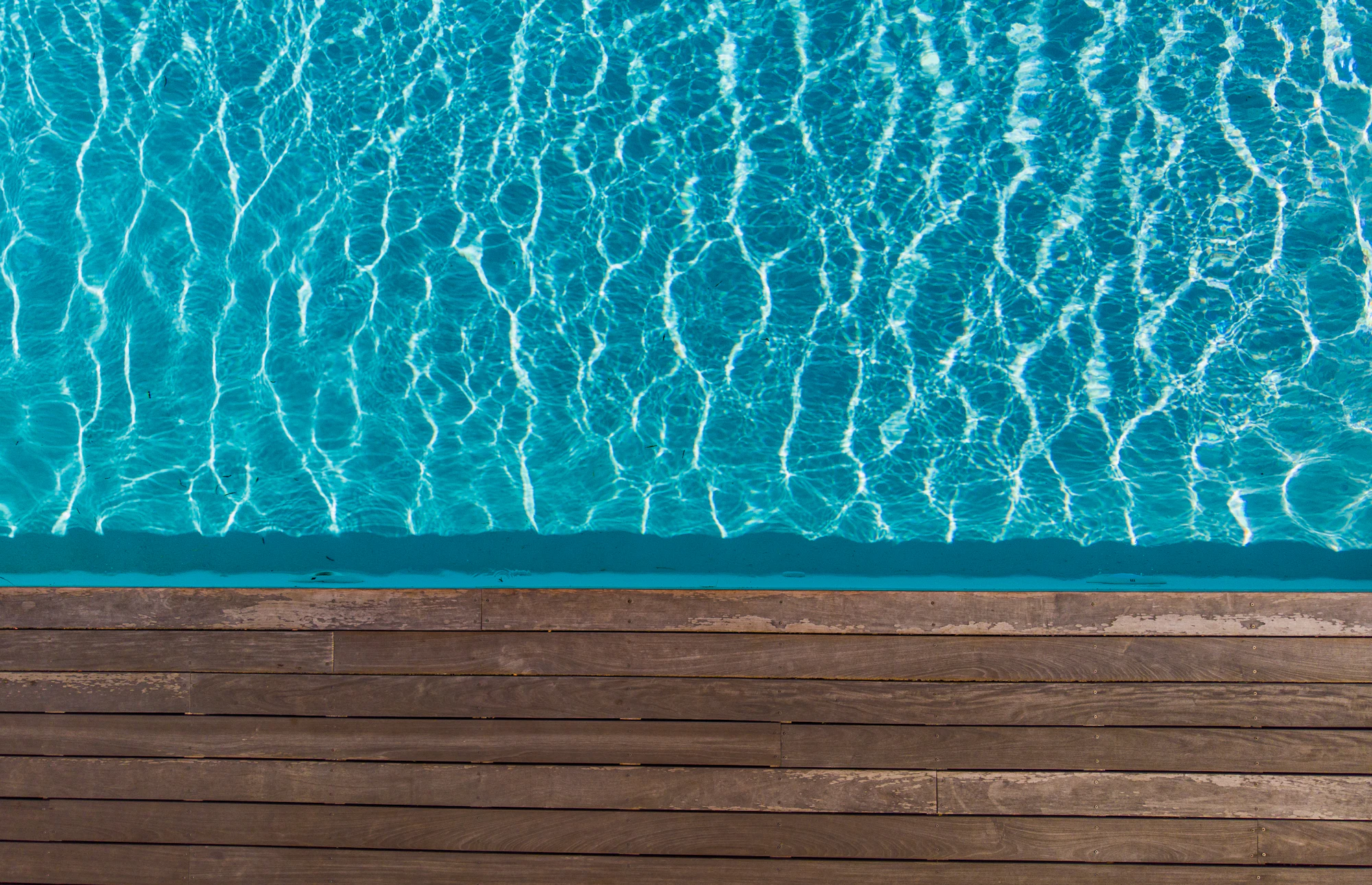 Overhead view of rippling turquoise pool water meeting a wooden deck