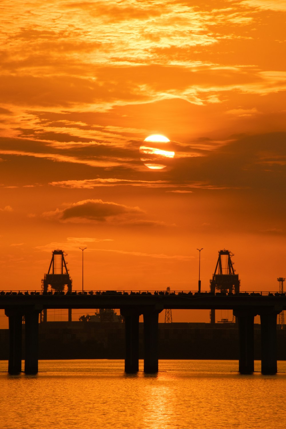 the sun is setting over the water with a bridge in the foreground