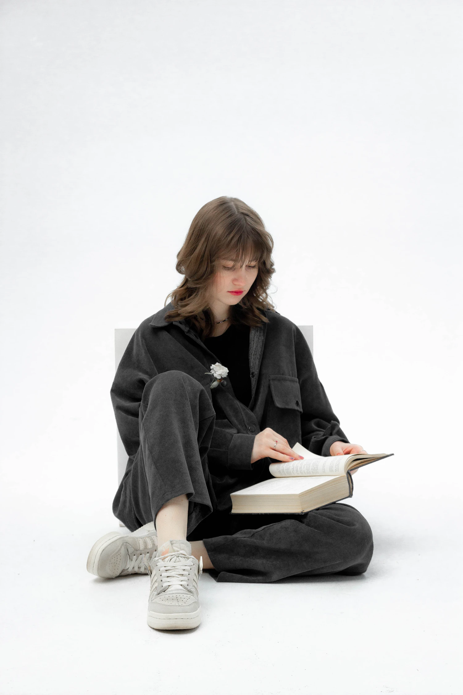 A figure sitting on the floor in a grey jacket and wide trousers, reading