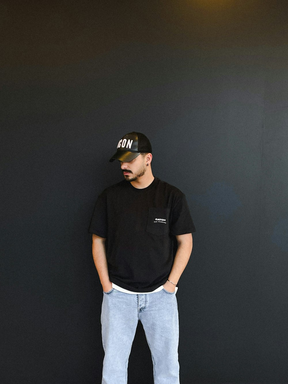 a man standing against a black wall wearing a hat