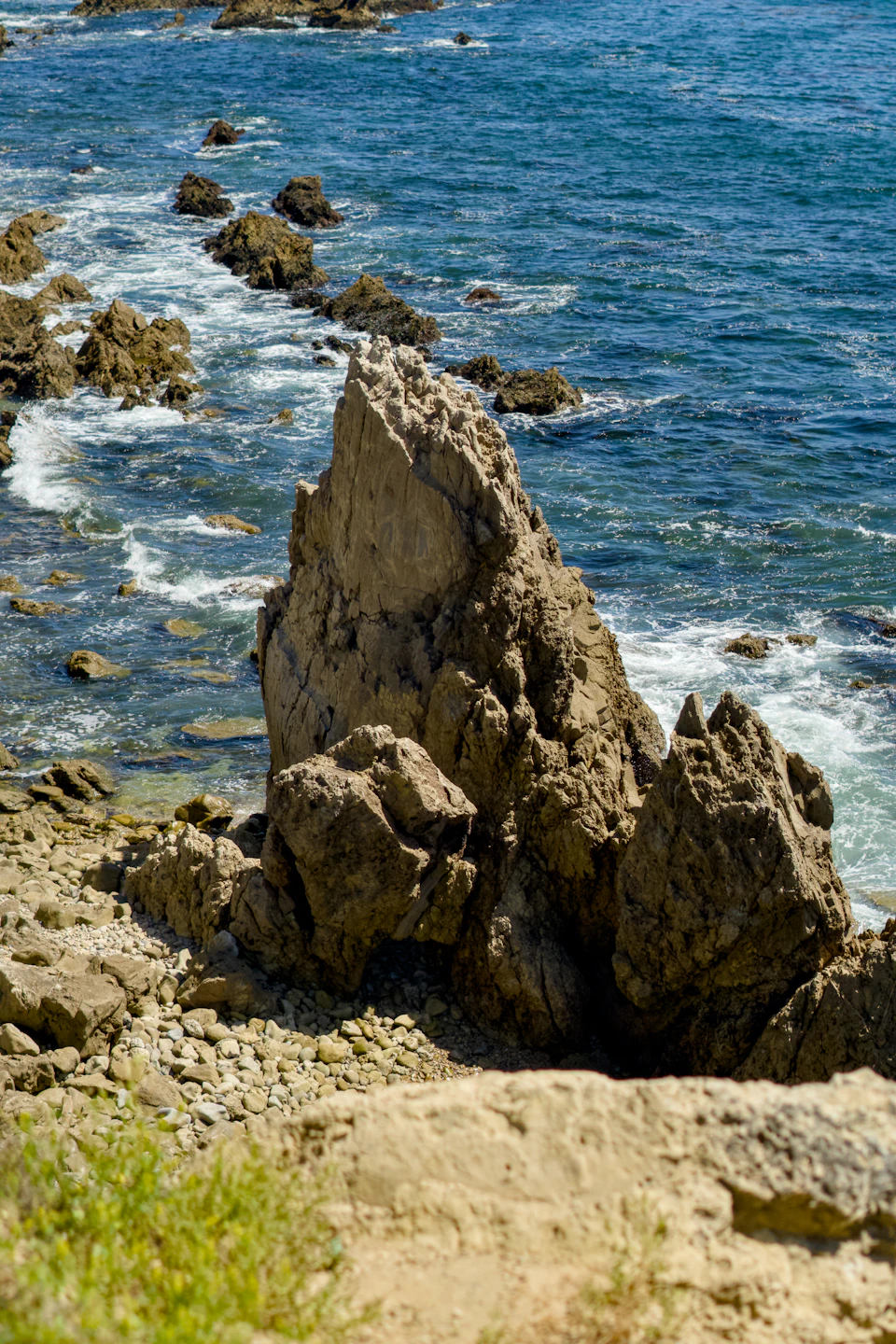 The image depicts a rocky coastline with a large, jagged rock formation on the left side and smaller rocks scattered around it. The water surrounding the rocks is a deep blue color, suggesting that the photo was taken during the day under clear skies. In the foreground, there are patches of green vegetation, adding a touch of life to the otherwise rugged landscape.