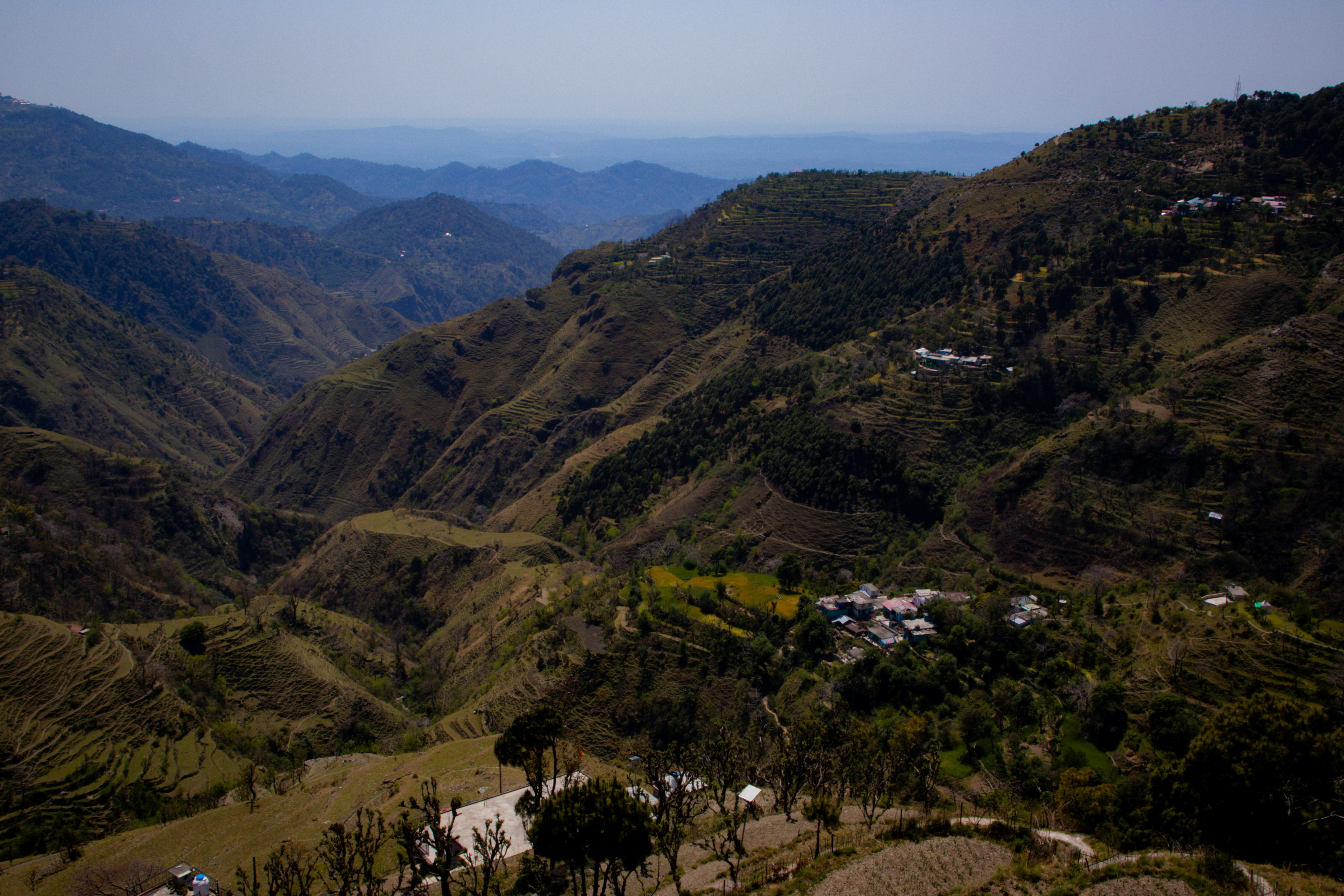 Rolling green highland plateau with terraced farmland and winding river valley visible in the distant hazy blue mountains