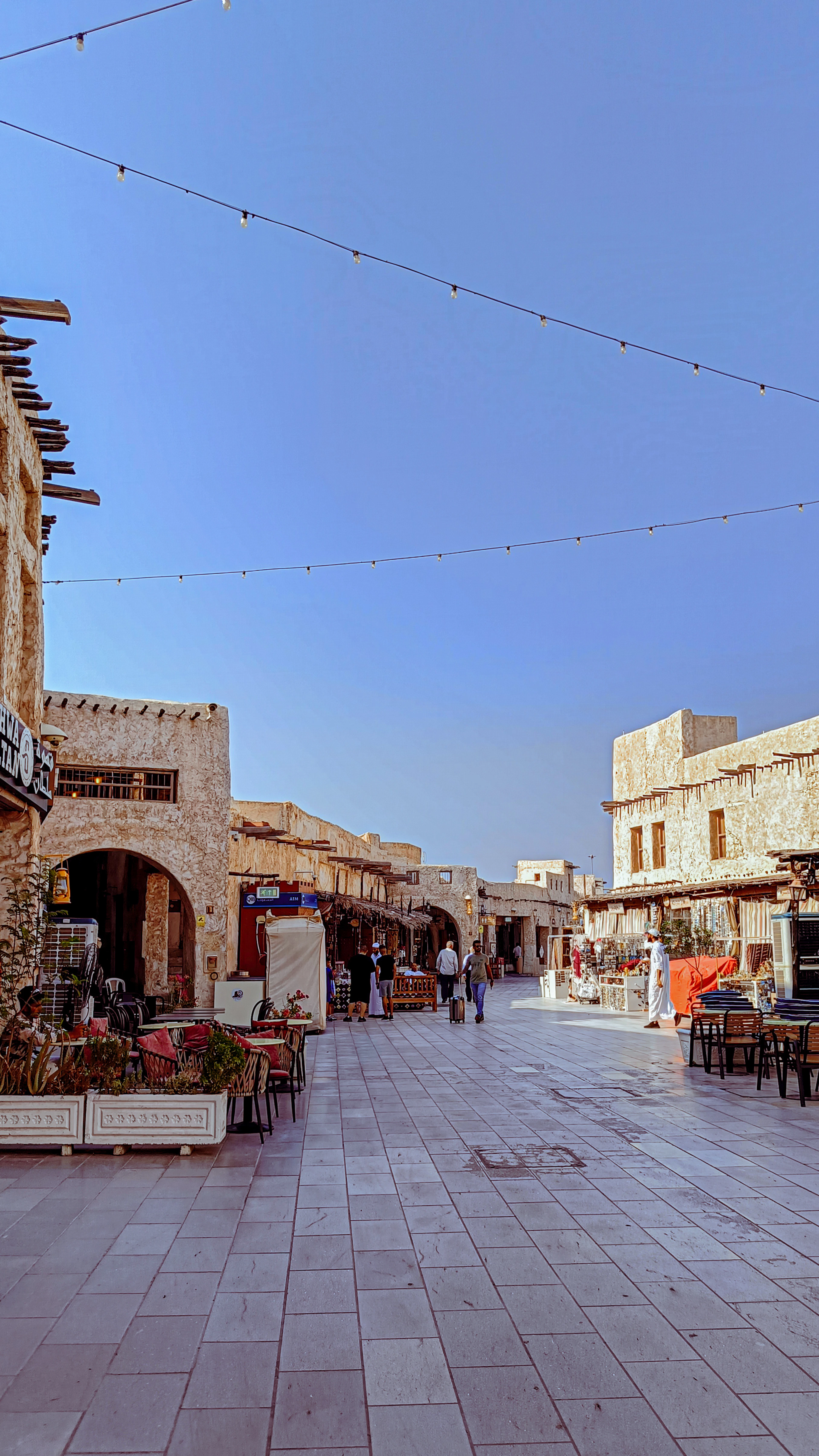 Souq Waqif in Doha in the last warm light of the day, its mud-rendered walls and protruding danchal roof poles lit gold, strings of festoon bulbs crossing the empty tiled lane and restaurant chairs and cushioned benches set out for the evening