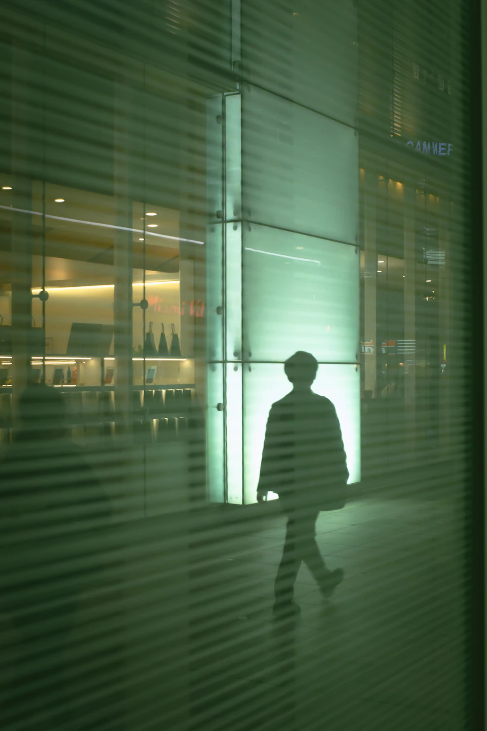 Person walks down sidewalk silhouetted against storefront with green glass and white frame