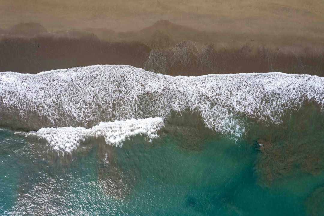 The aerial shot shows waves gently lapping a sandy shore, creating foam and intricate patterns in the sea. This image of the tranquil beach from above showcases the serene interface between ocean and land, highlighting the calming hues and textures of the natural seascape.