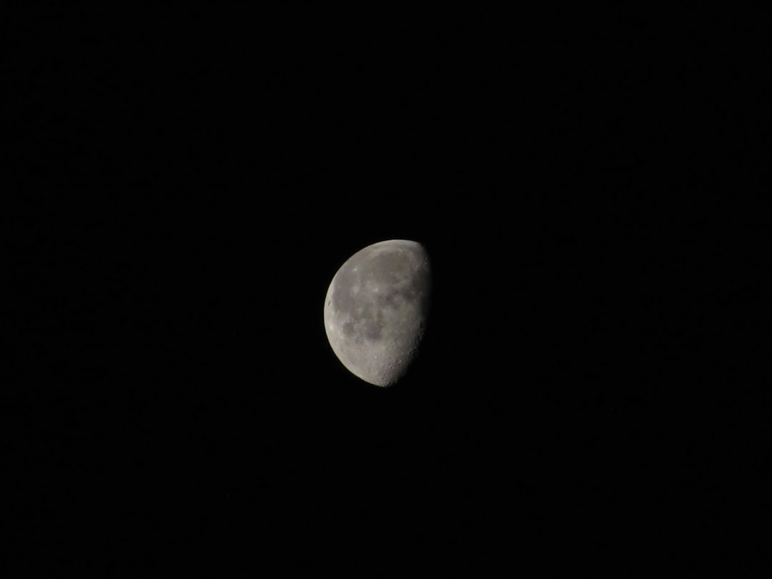 A night sky photo featuring a half moon, its illuminated surface a bright white against the dark blue background. The moon's craters and ridges are visible in detail.