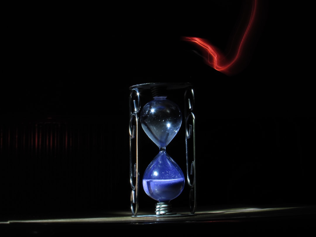 A close-up photo of a hourglass resting on a wooden table. The hourglass has a glass top and bottom section connected by a thin neck filled with white sand. Some of the sand has trickled down to the bottom section, creating an artistic light drawing through the center.