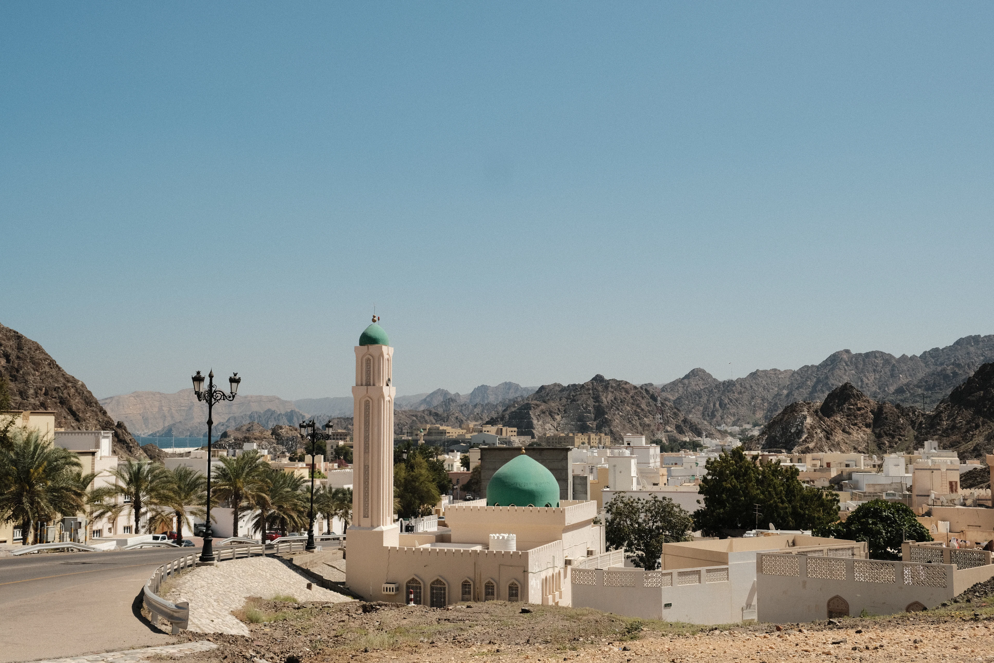 A mosque with a green dome and minaret in a coastal district of Muscat, bare mountain ridges beyond, Oman