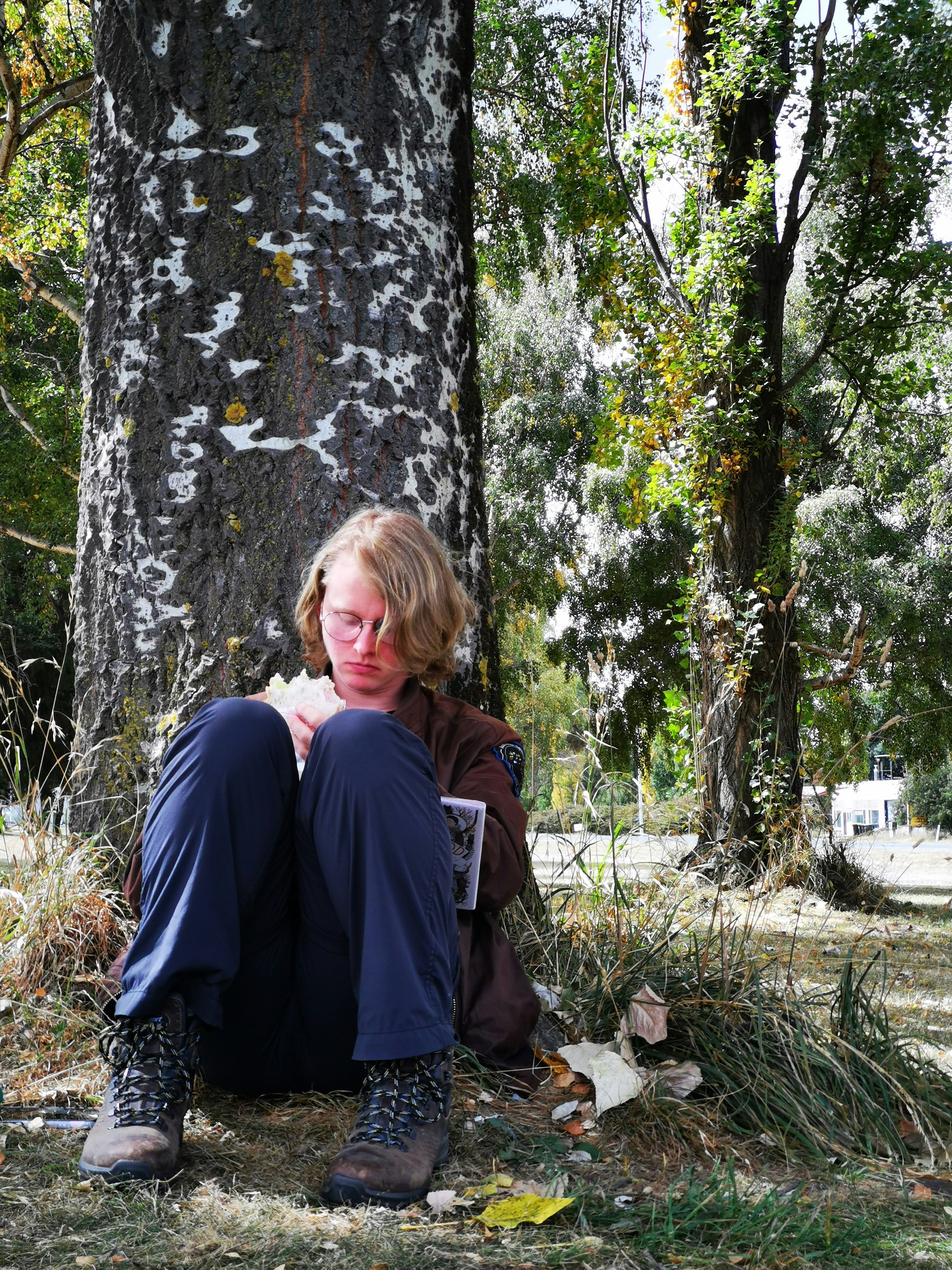 A person sitting against a tree trunk, reading a book.