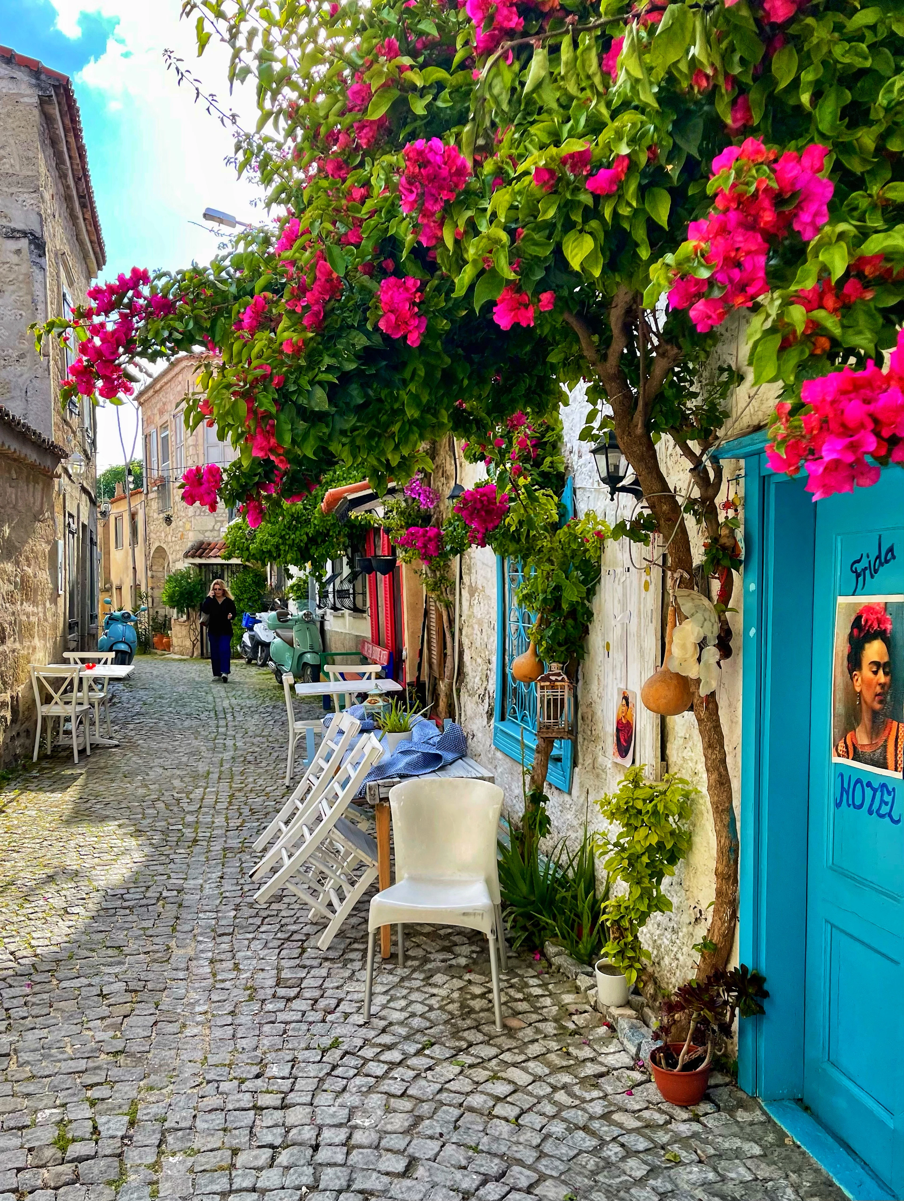 A cobbled lane in Alacati near Izmir, a canopy of magenta bougainvillea overhanging pale stone houses, cafe tables and chairs set out along the paving and a turquoise-painted hotel door at the right