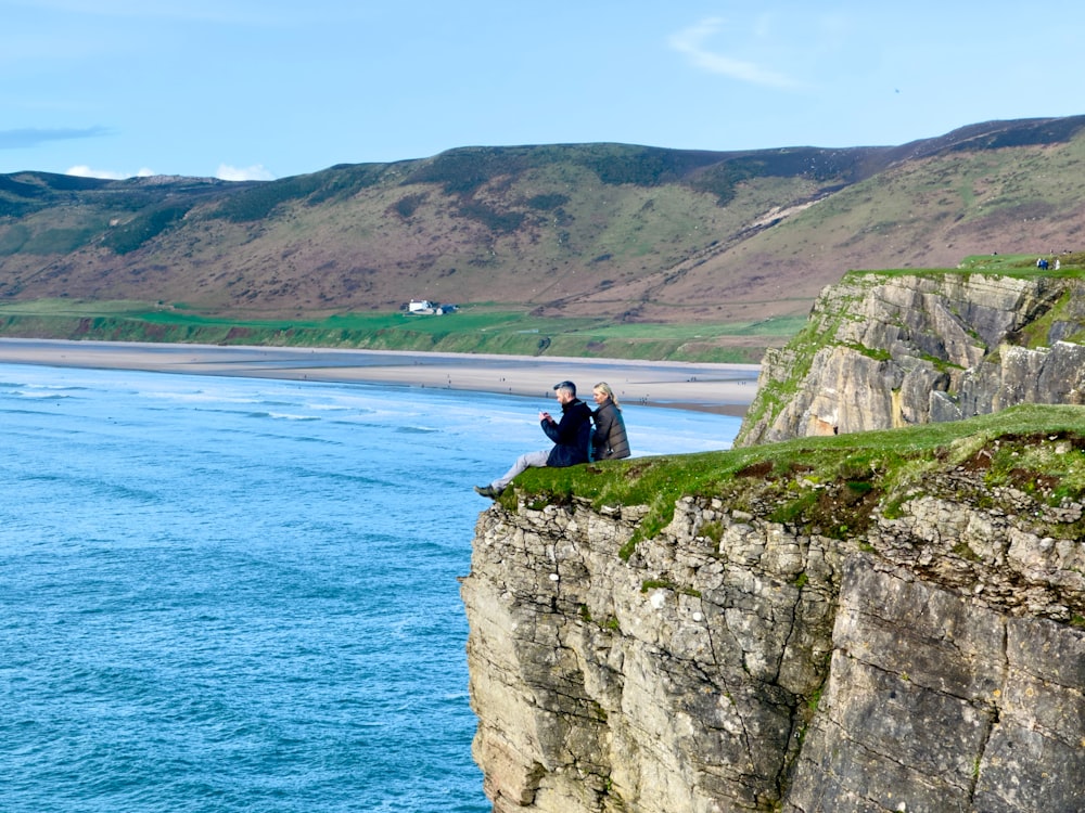 two people sitting on a cliff overlooking a body of water