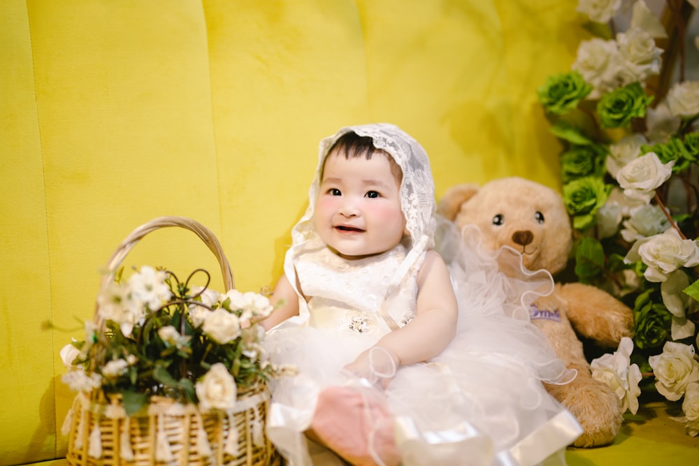 a baby in a white dress sitting next to a teddy bear