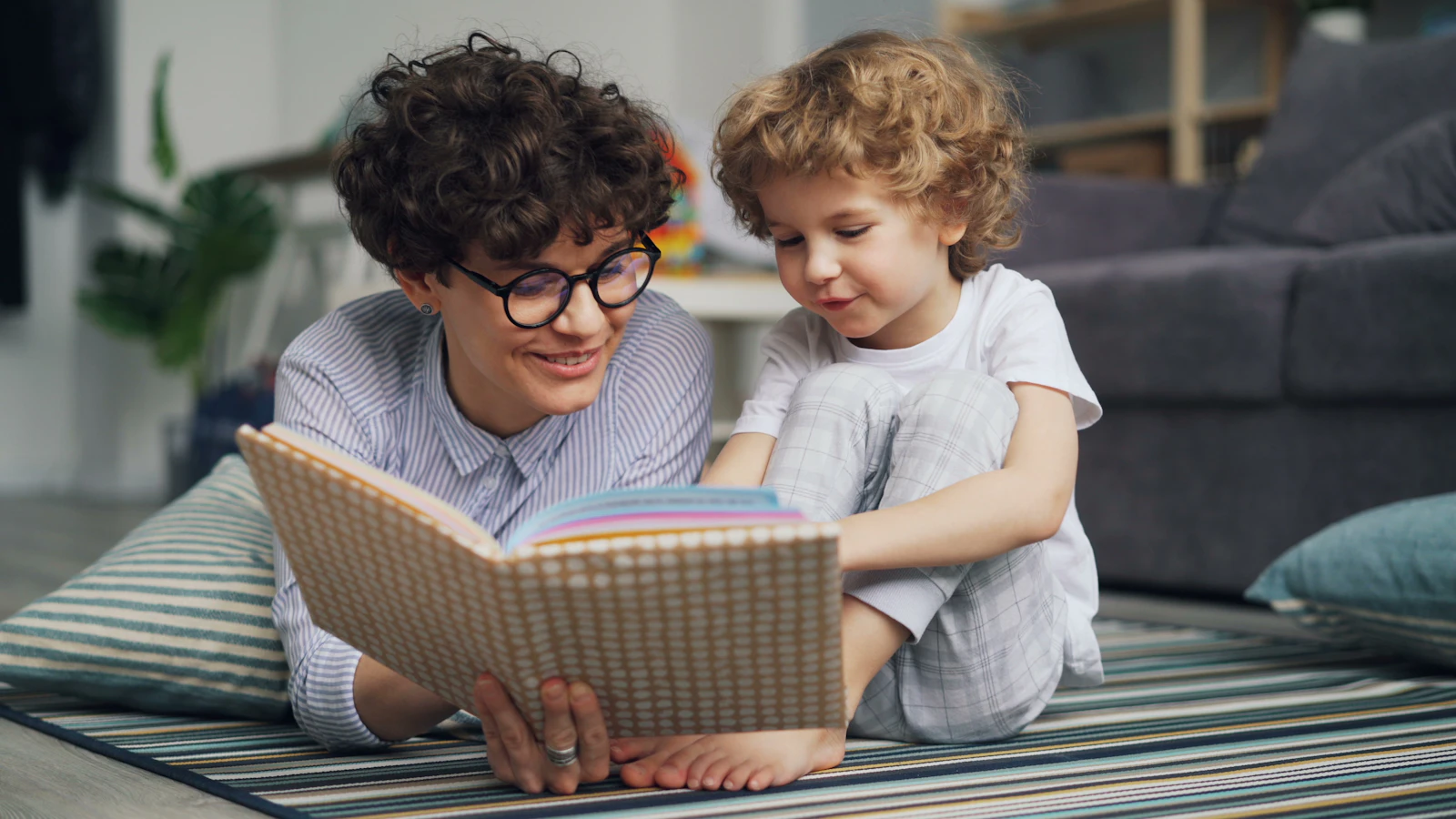 An adult and a young child lying on a rug together looking at an open picture book