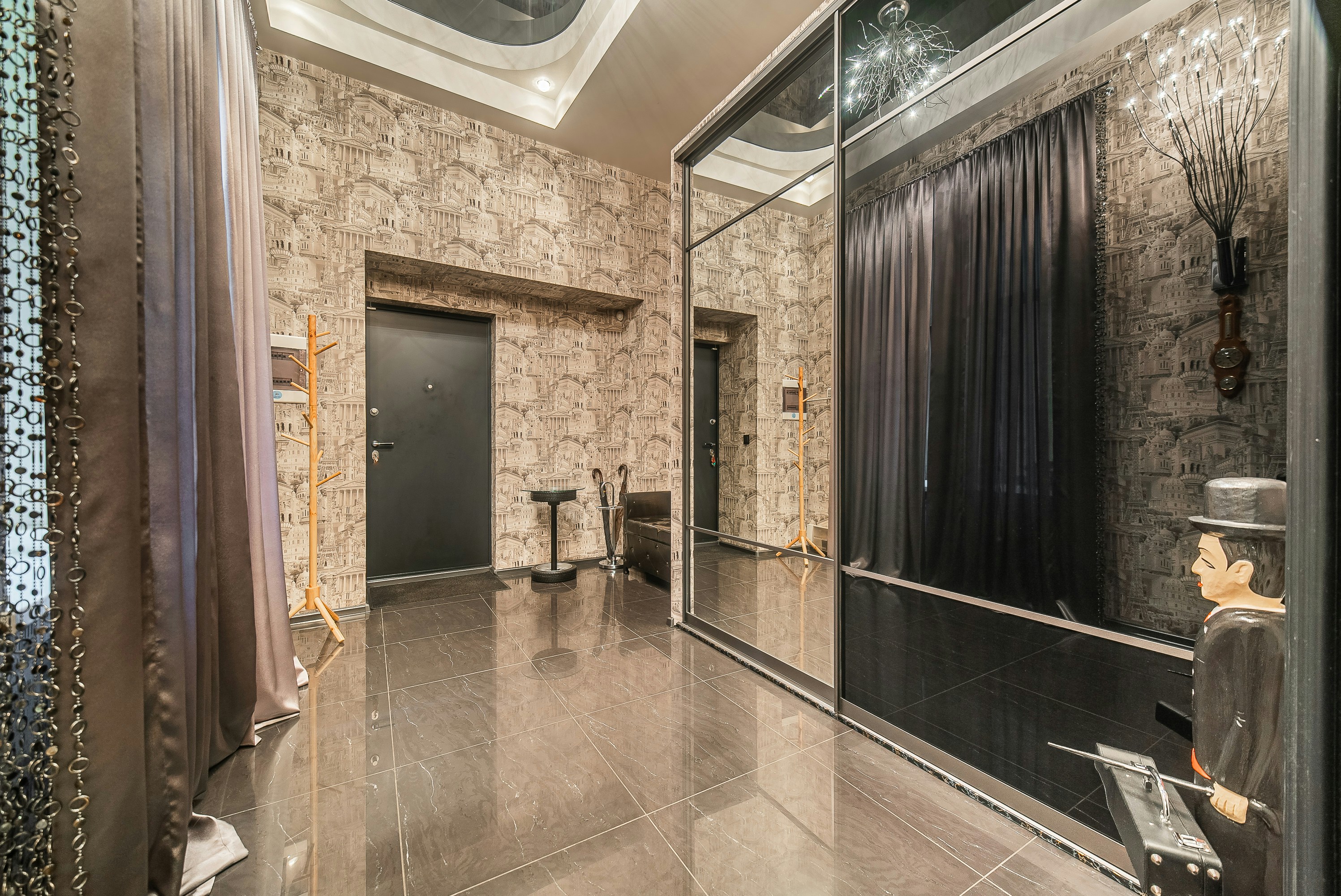 Modern hallway featuring ornate patterned wallpaper, dark tiled floor, a black door, and a large mirrored wardrobe with quirky decor.