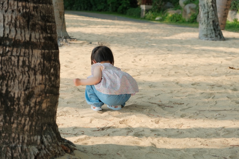 a little girl sitting in the sand next to a tree
