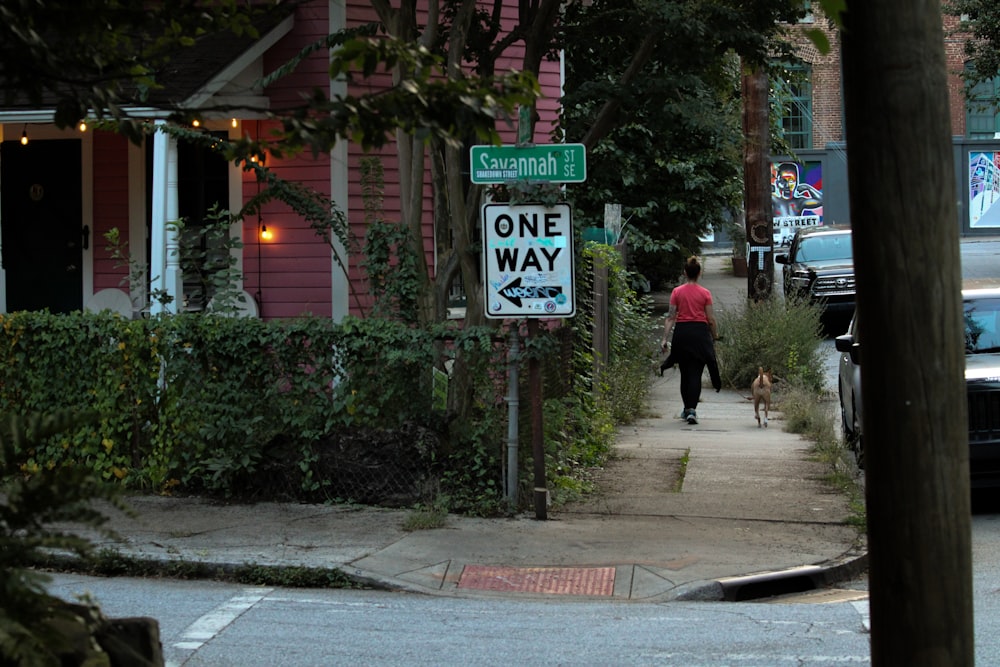 a woman walking a dog down a sidewalk next to a one way sign
