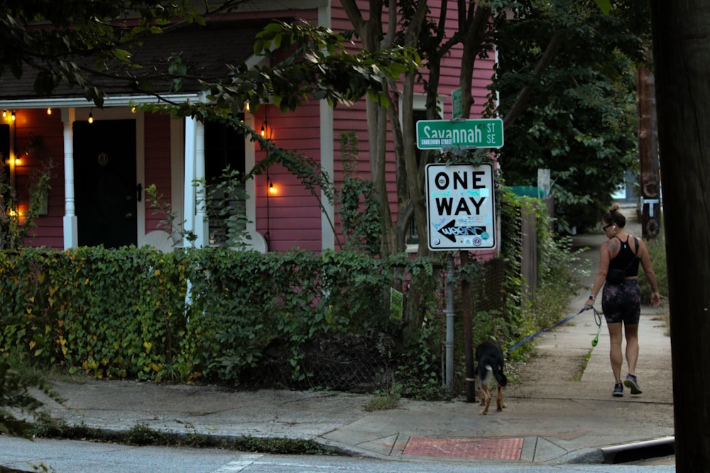 a woman walking a dog down a sidewalk next to a one way sign