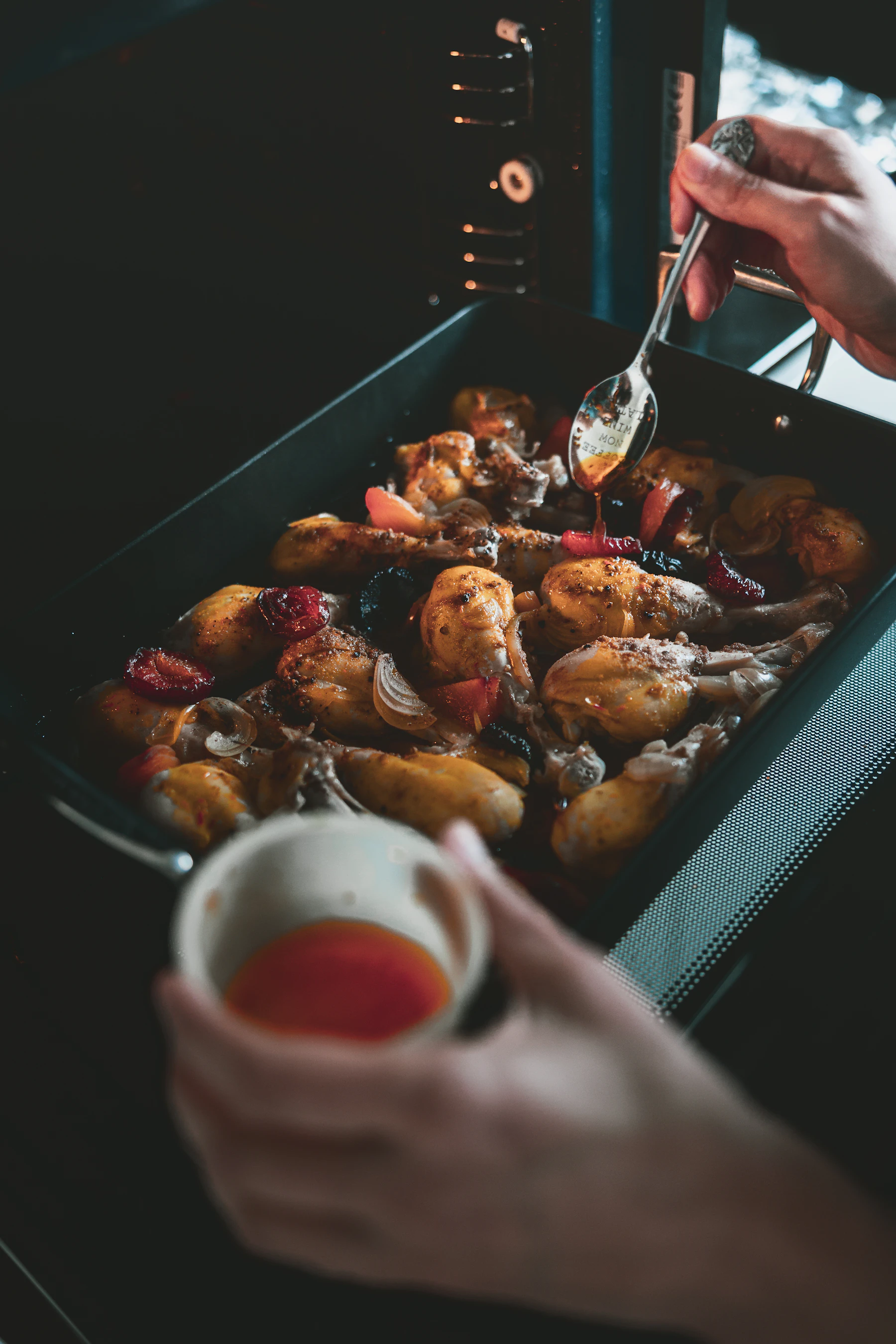 A cook spooning pan juices over chicken drumsticks in a roasting pan at the open oven door