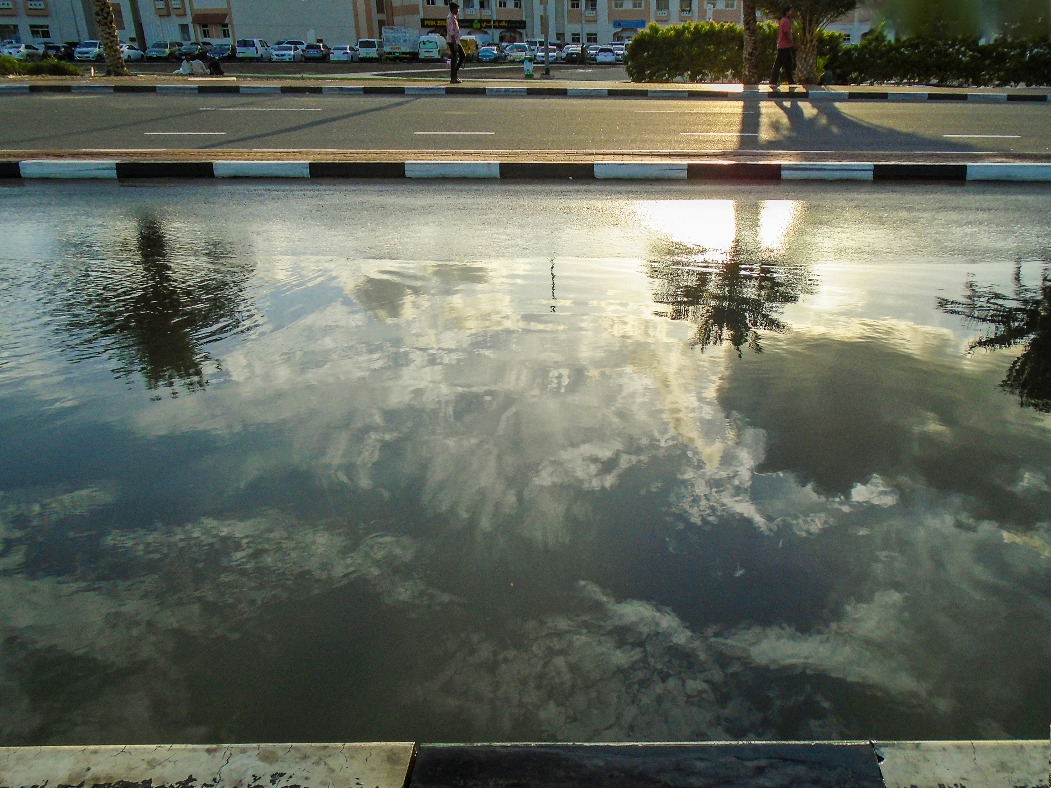 Reflective urban pool along a street with a curb and buildings in the background; the water captures clouds and sky like a still mirror.