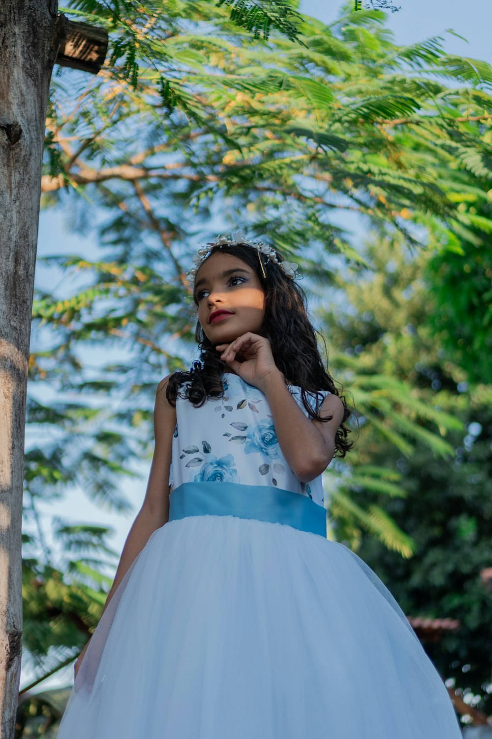 a little girl in a dress standing next to a tree