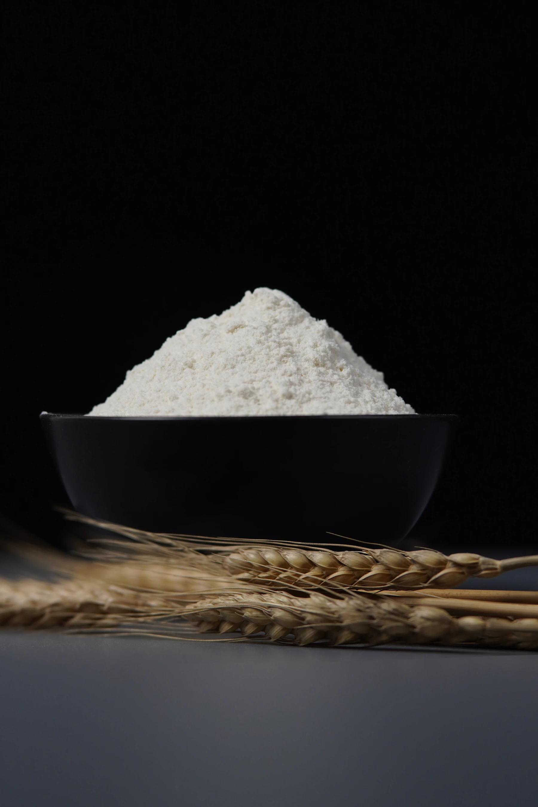 A heaped black bowl of flour with dried wheat ears lying in front of it on a dark backdrop