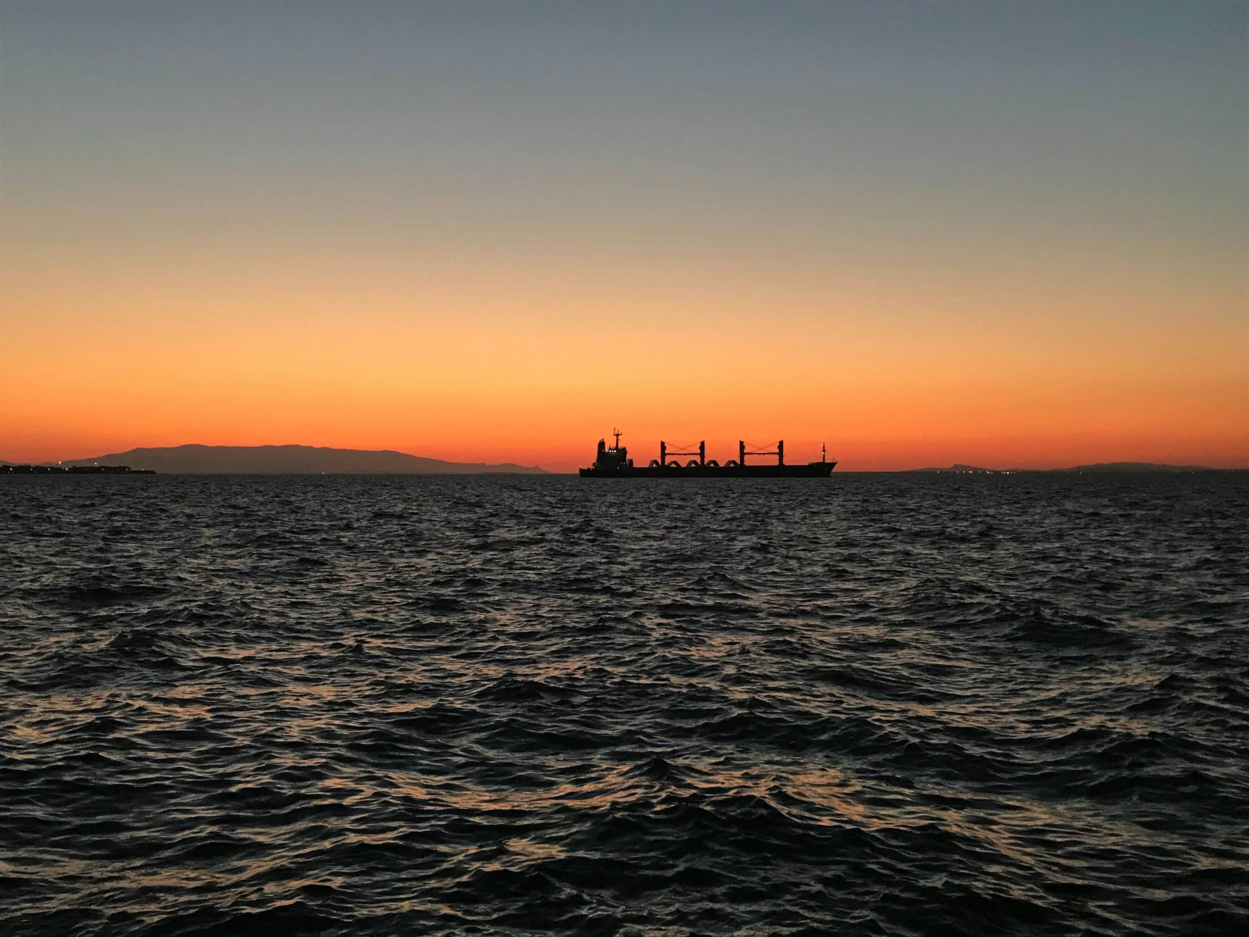 Massive cargo vessel at golden hour on dark ocean, dramatic low-key lighting with amber horizon glow